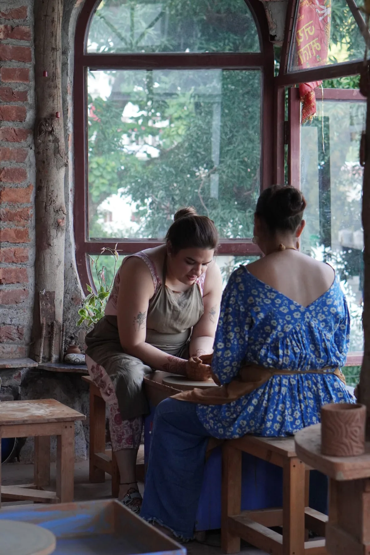 Two guests shaping clay at the wheel beside the arched studio window